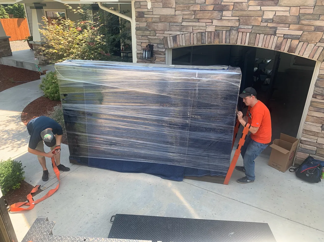 Two workers moving a large wrapped object on a driveway in front of a stone house