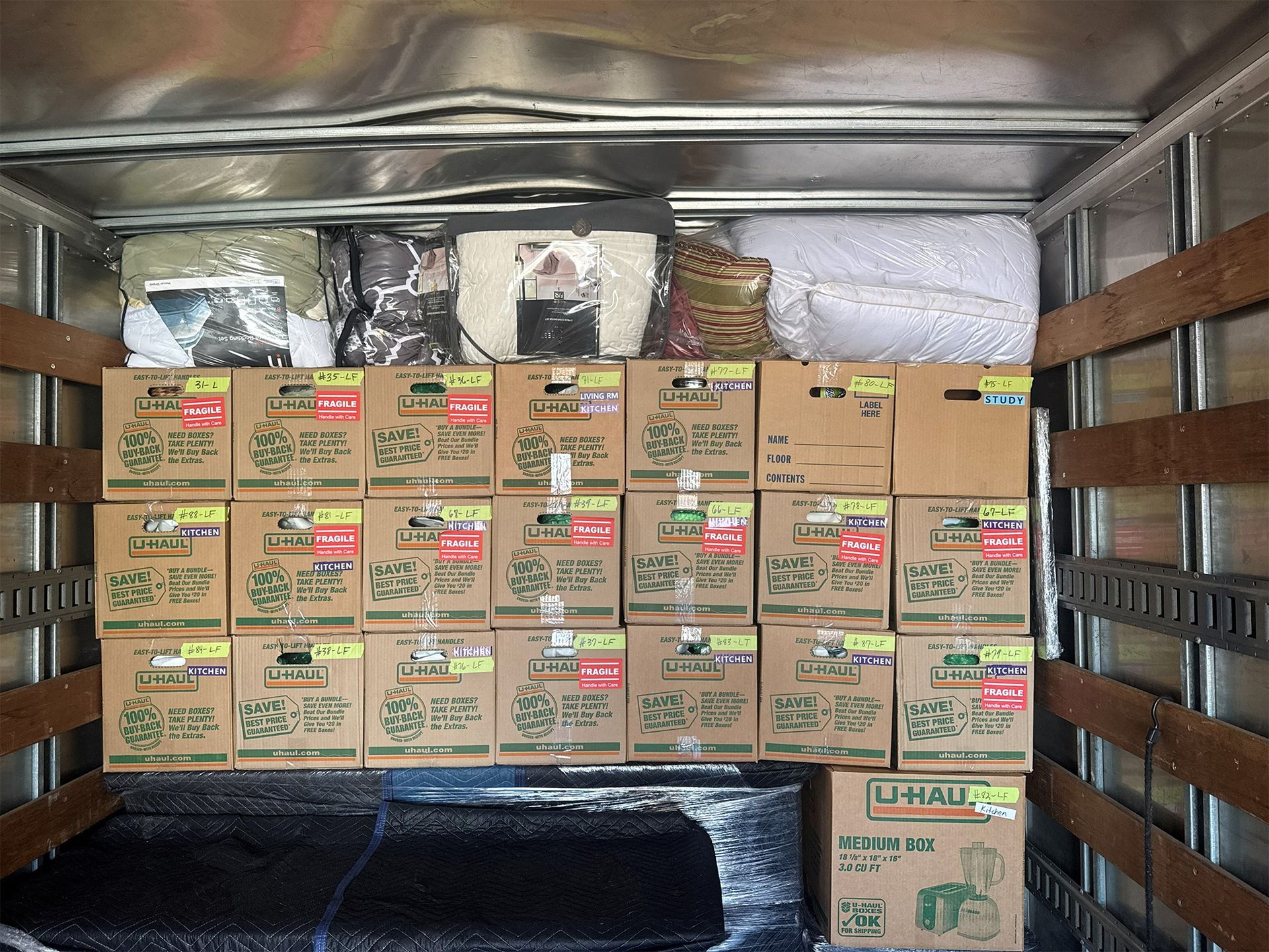 Packed cardboard boxes stacked inside a cargo trailer, with metal walls and a black floor mat.