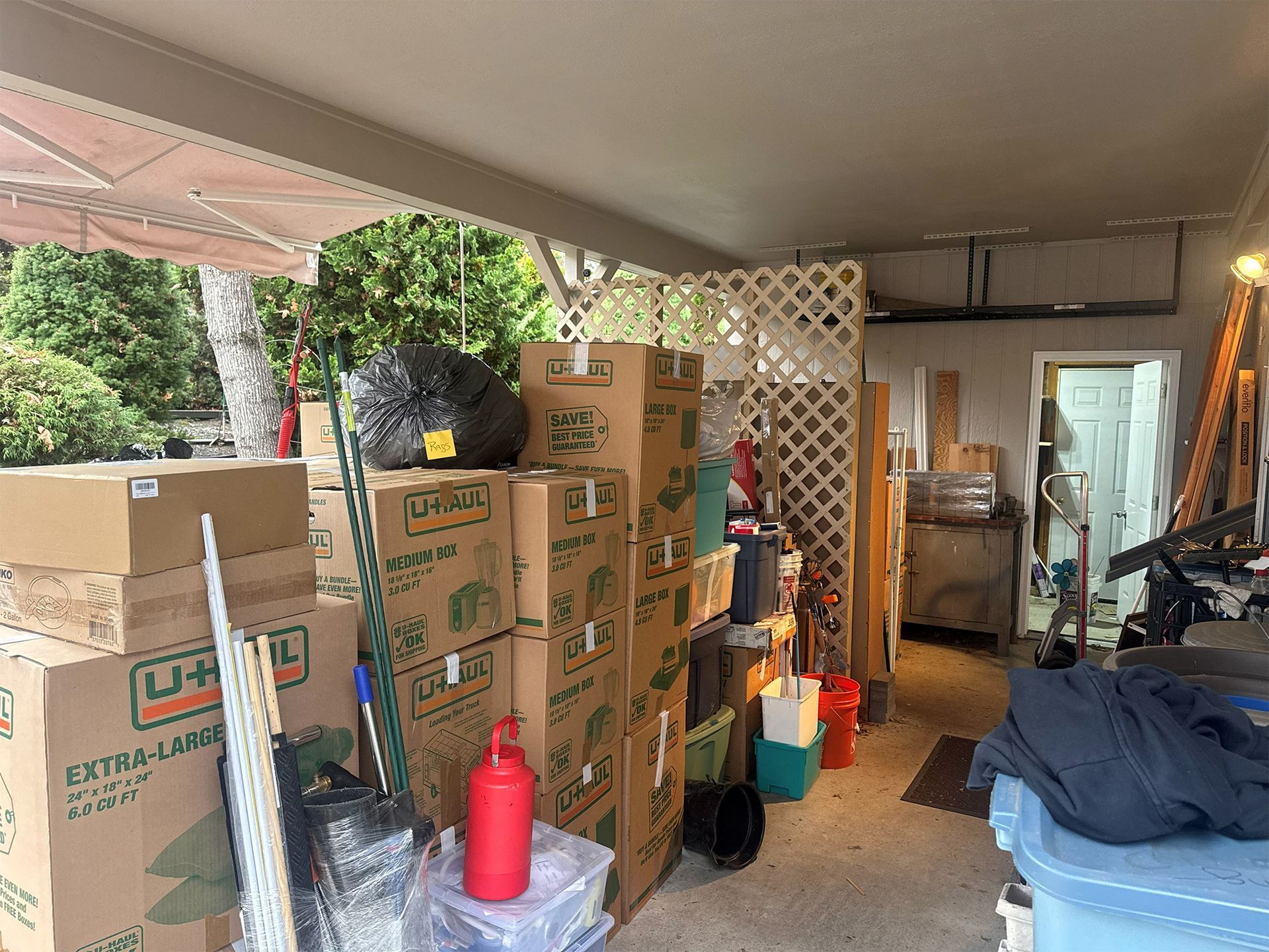 Packed garage with stacked moving boxes, a red gas can, and outdoor equipment under a covered patio.