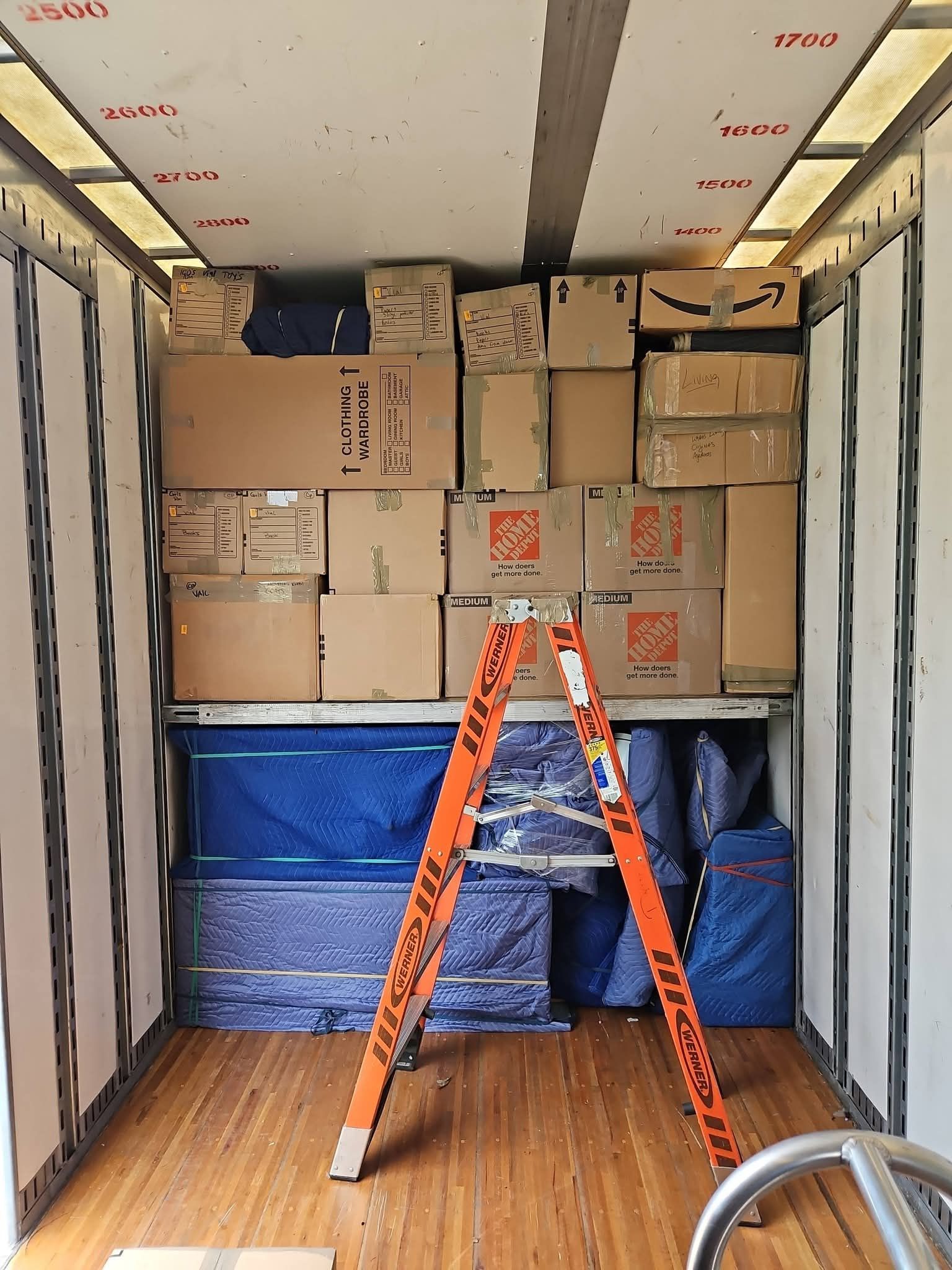 Moving truck interior stacked with boxes and blue wrapped items, with an orange ladder in front.