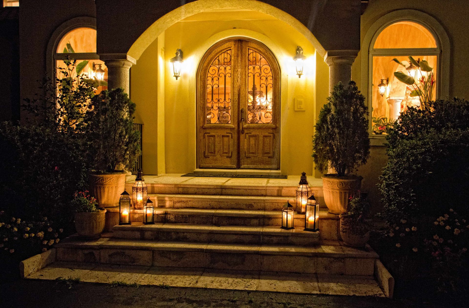 Lit entryway of a home at night. Steps lead to ornate double doors, flanked by lanterns and potted plants. Yellow glow.