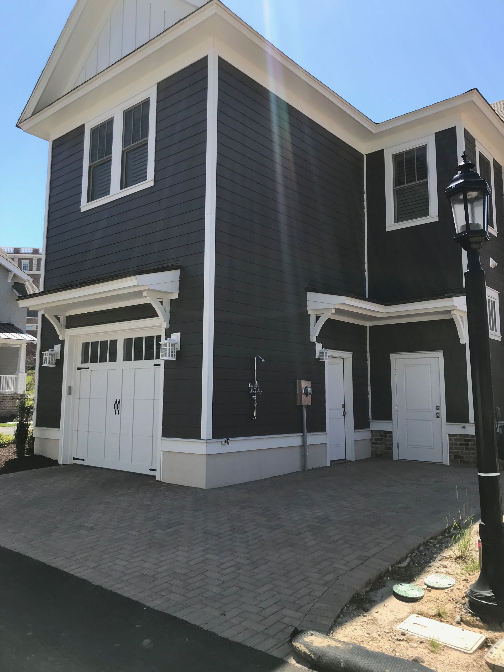 Two-story house with dark gray siding, white trim, and a brick driveway. A garage door and two doors are visible.
