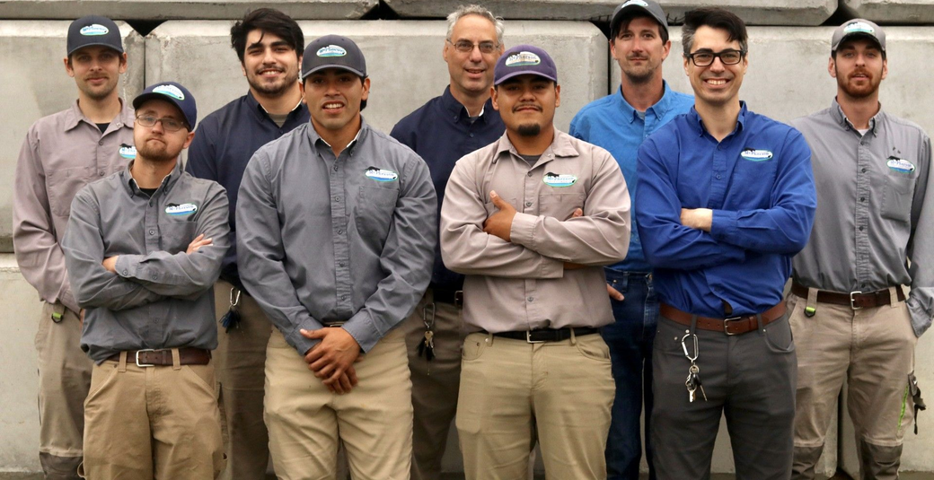 Seven workers standing shoulder to shoulder in gray and blue uniforms against a concrete wall