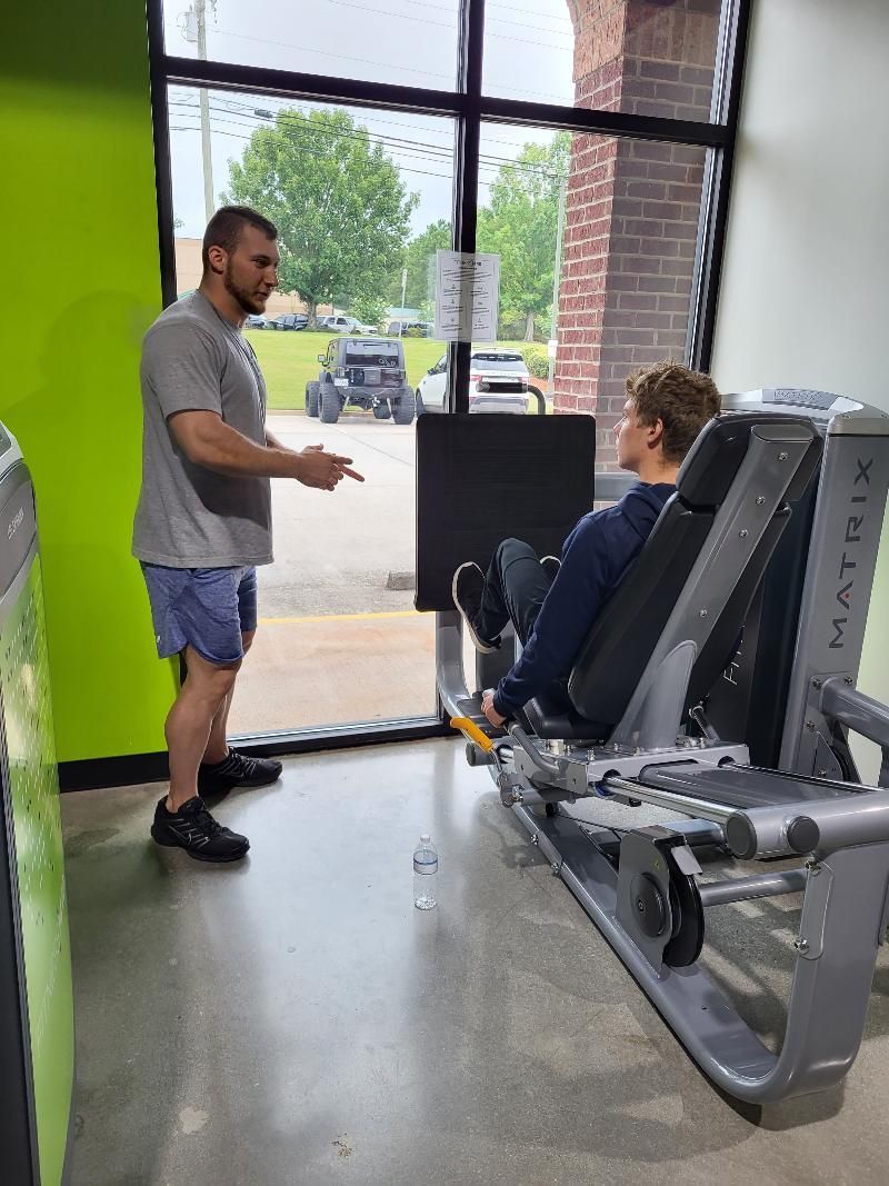 A man is standing next to a woman sitting on a machine in a gym.
