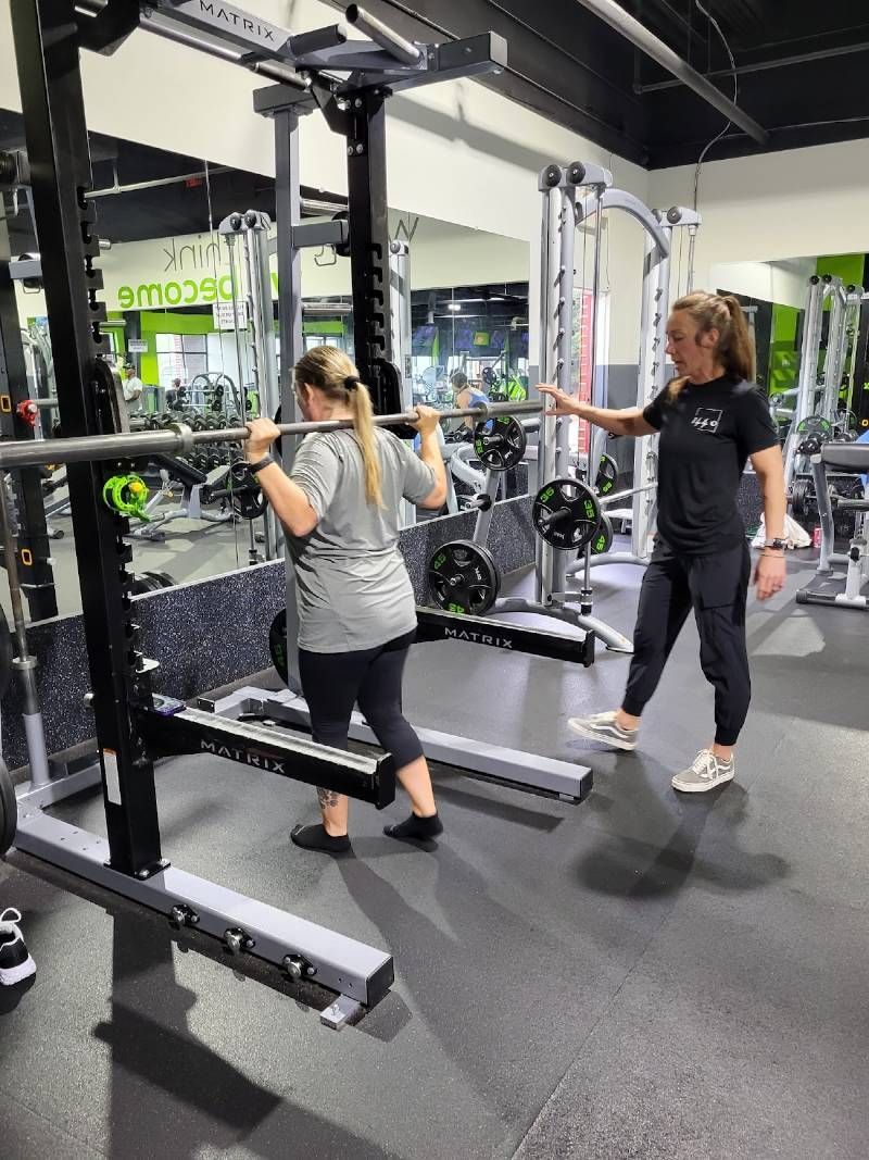 A woman is squatting with a barbell in a gym while another woman watches.