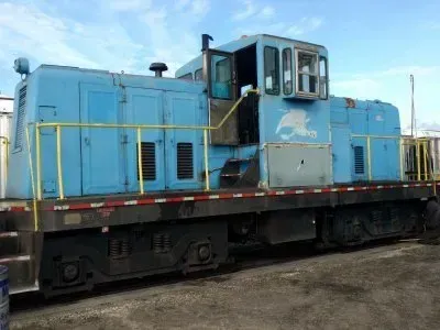 Blue diesel locomotive on tracks, side view, with yellow railings and a cloudy sky background