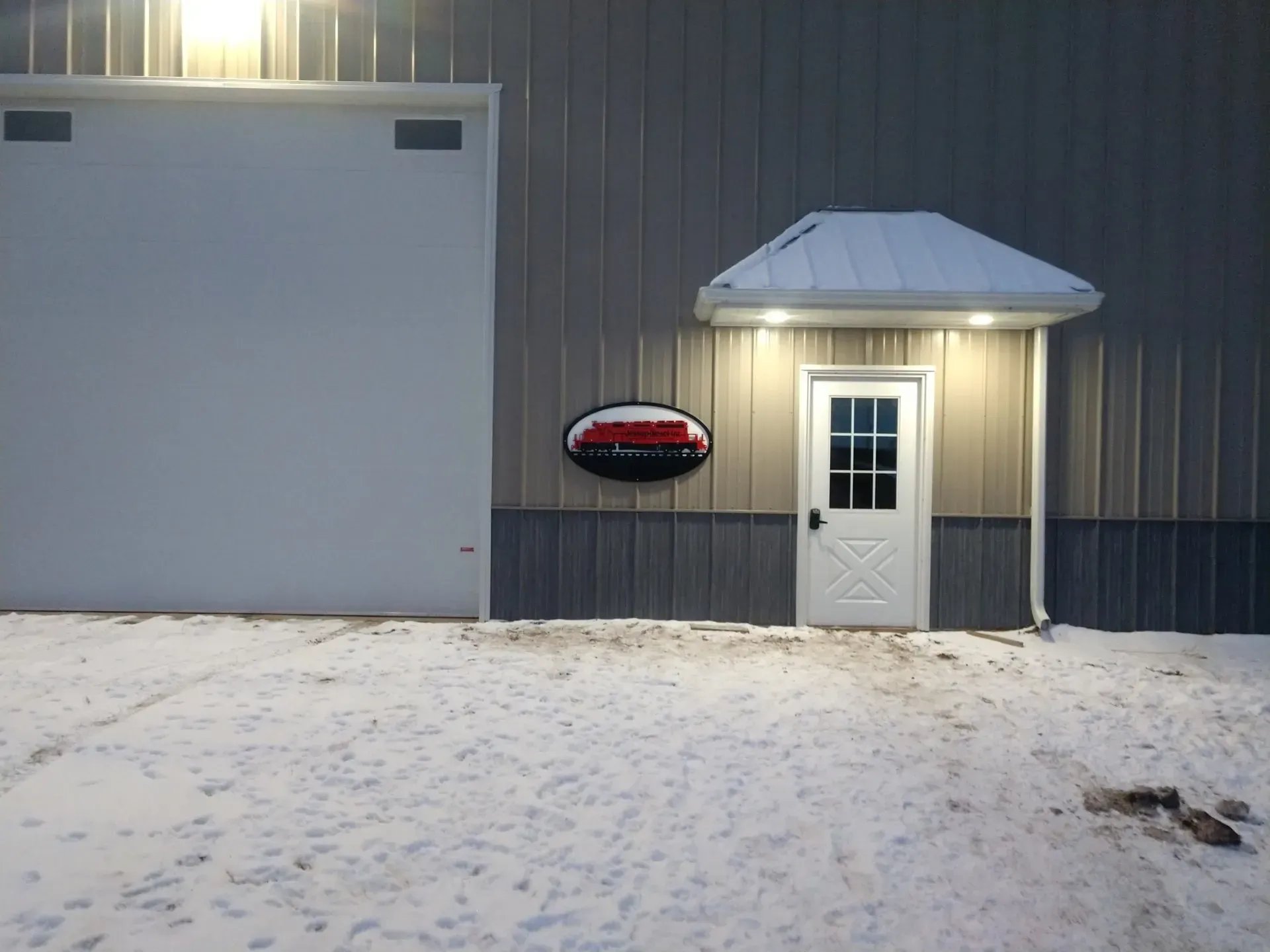 Snow-covered building exterior with a lit white door, small awning, and sign on gray siding.