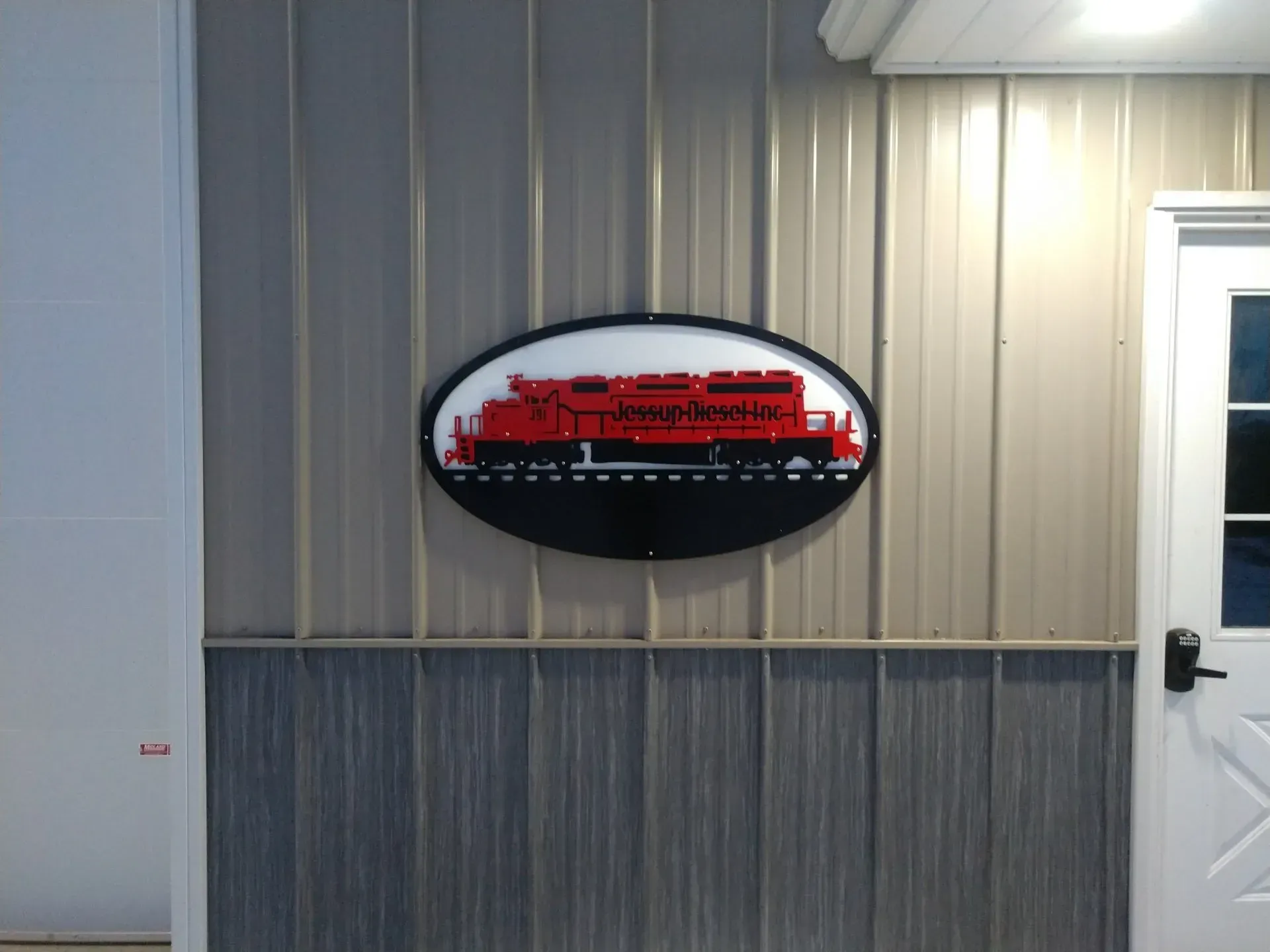 Oval red-and-black sign on a beige metal building wall beside a white door