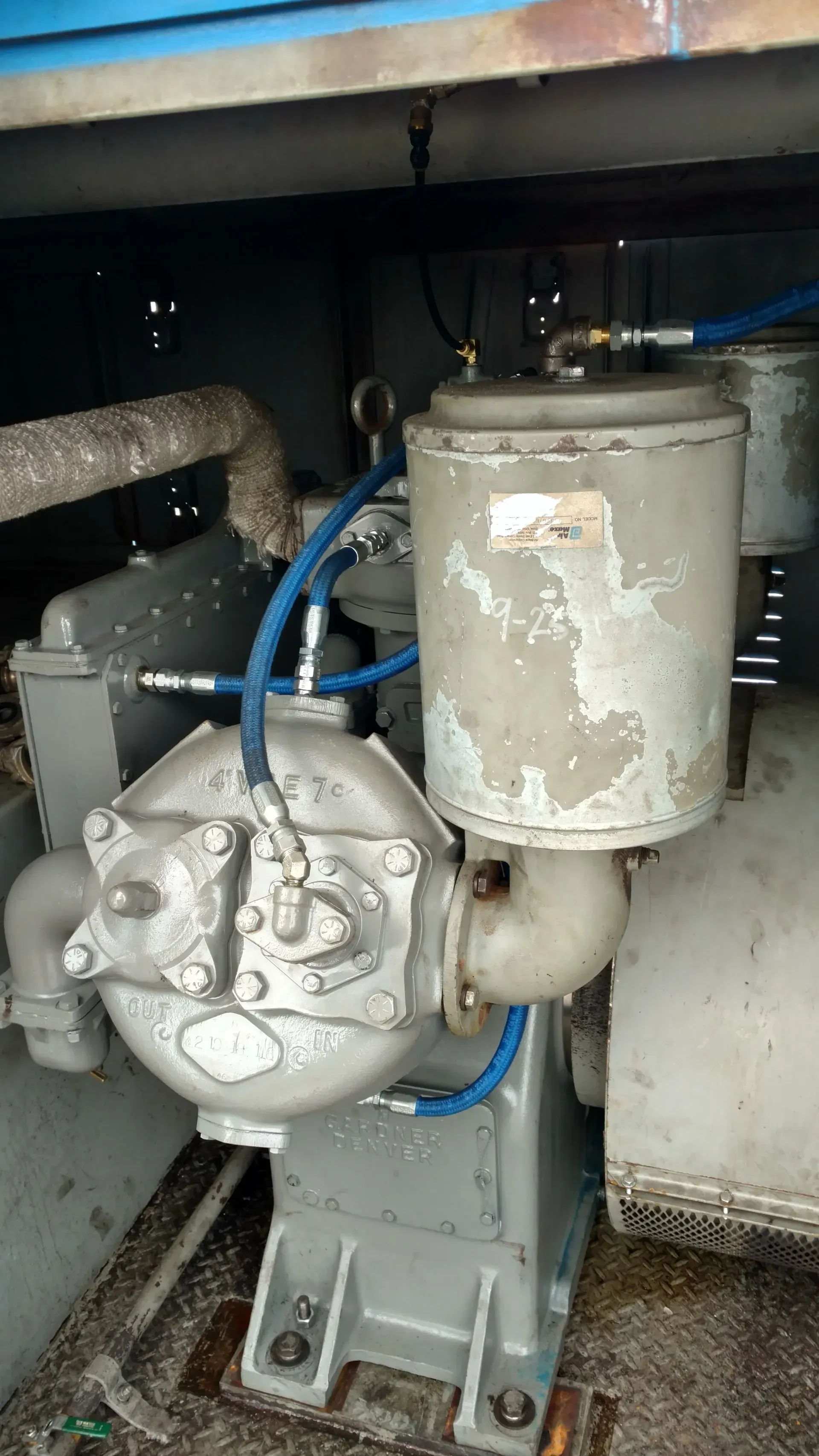 Grey industrial pump with blue hoses and a worn cylindrical tank mounted under a metal structure