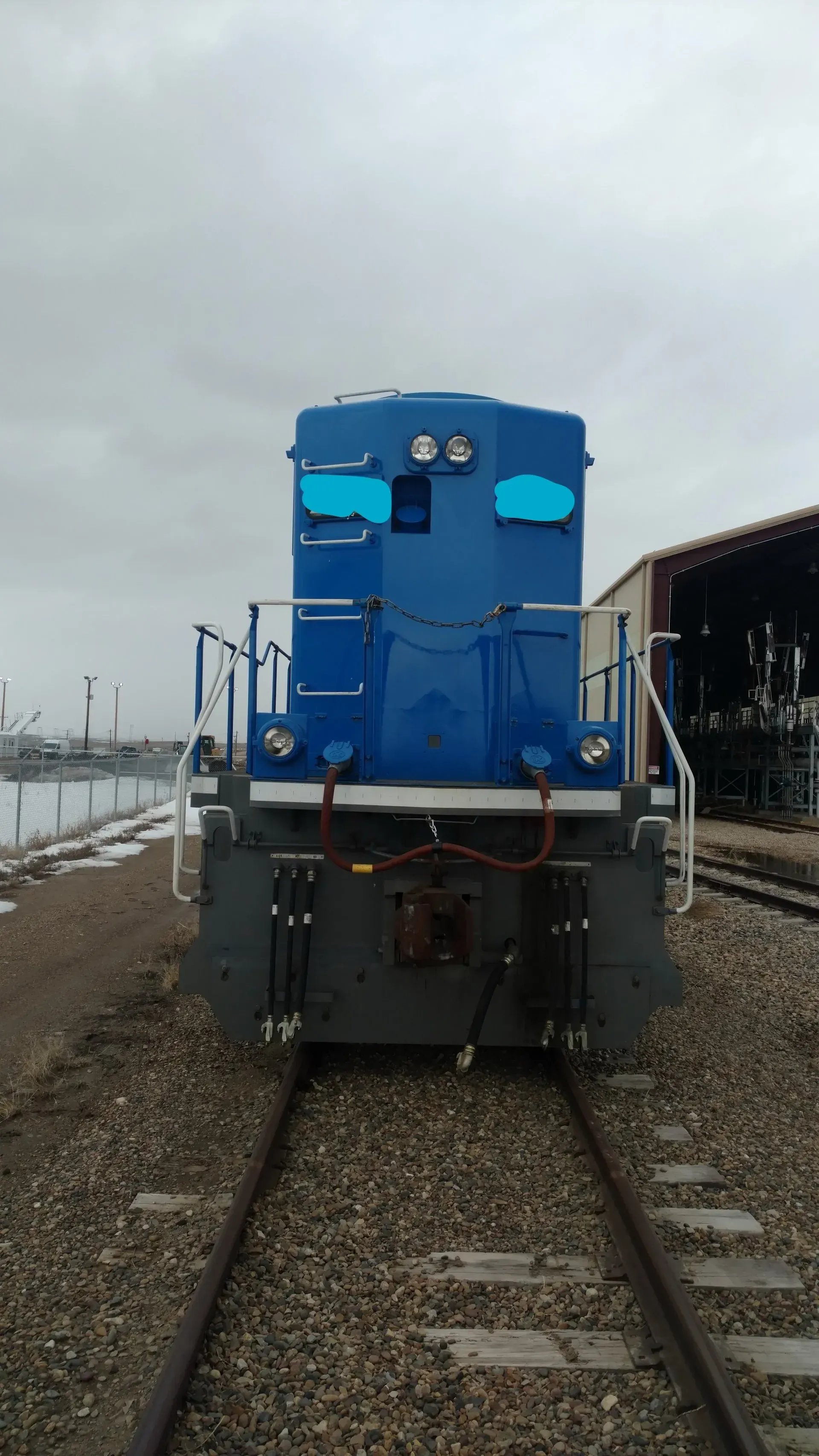 Blue locomotive facing forward on railroad tracks at a rail yard under an overcast sky