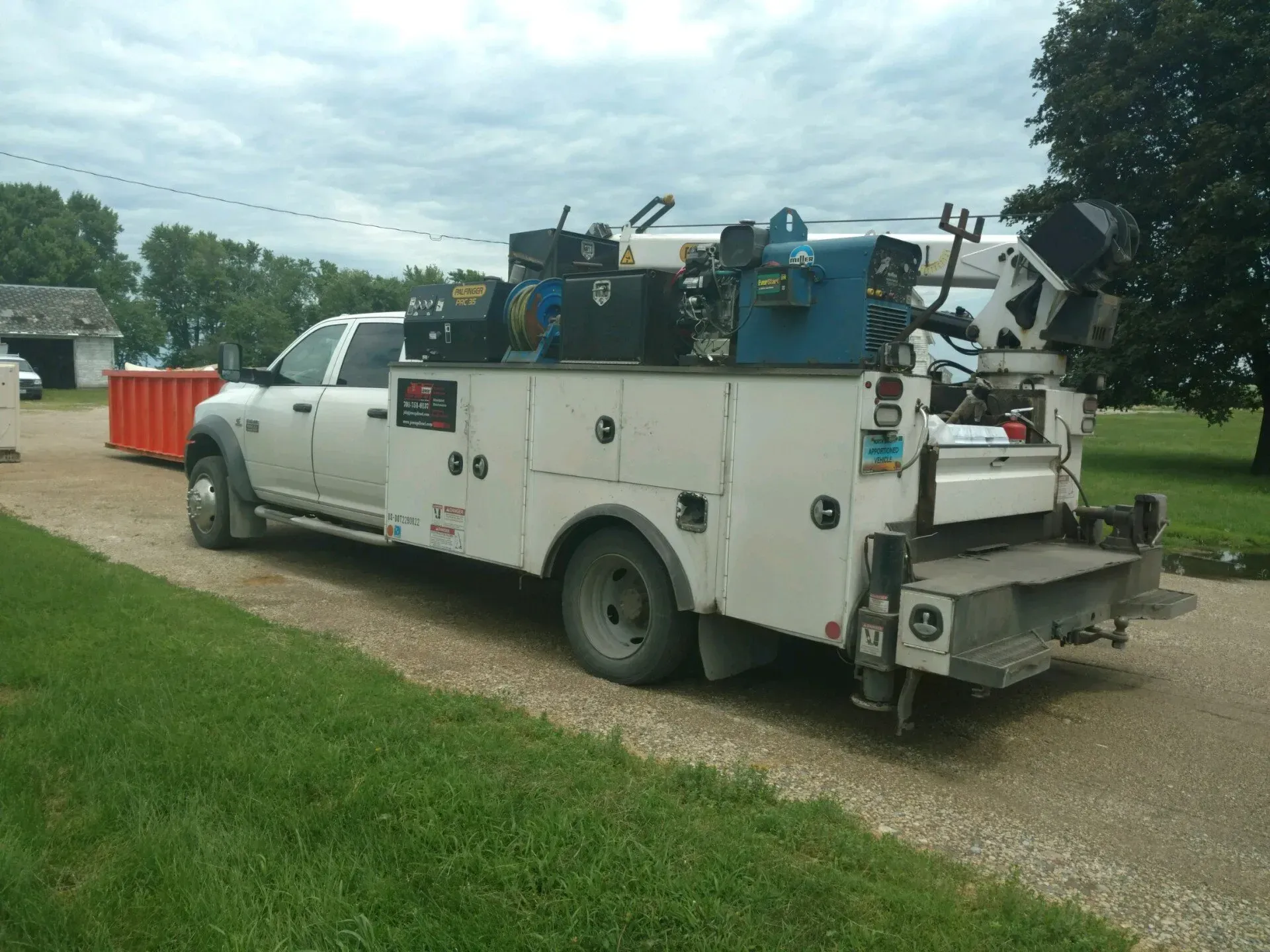 White utility truck with equipment parked on a gravel road beside a grassy field under cloudy skies