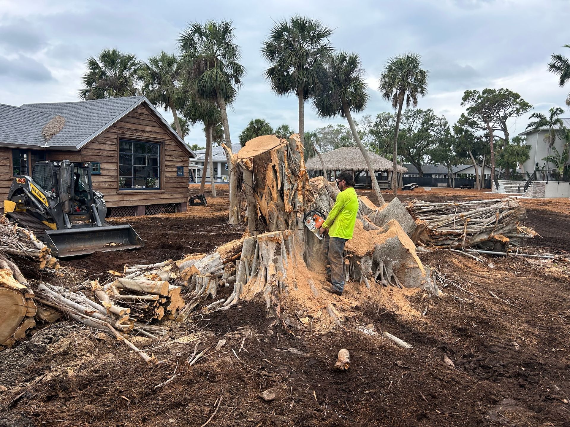 Man in yellow vest using chainsaw on large tree stump near house and machinery.