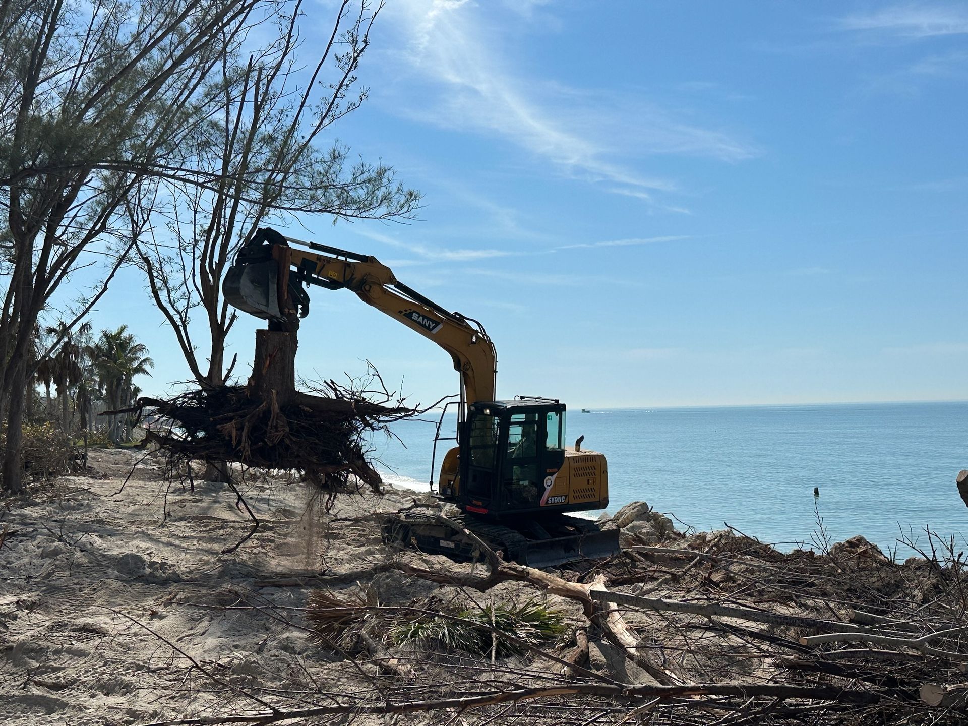 Yellow excavator removing tree roots from a beach next to the ocean under a blue sky.