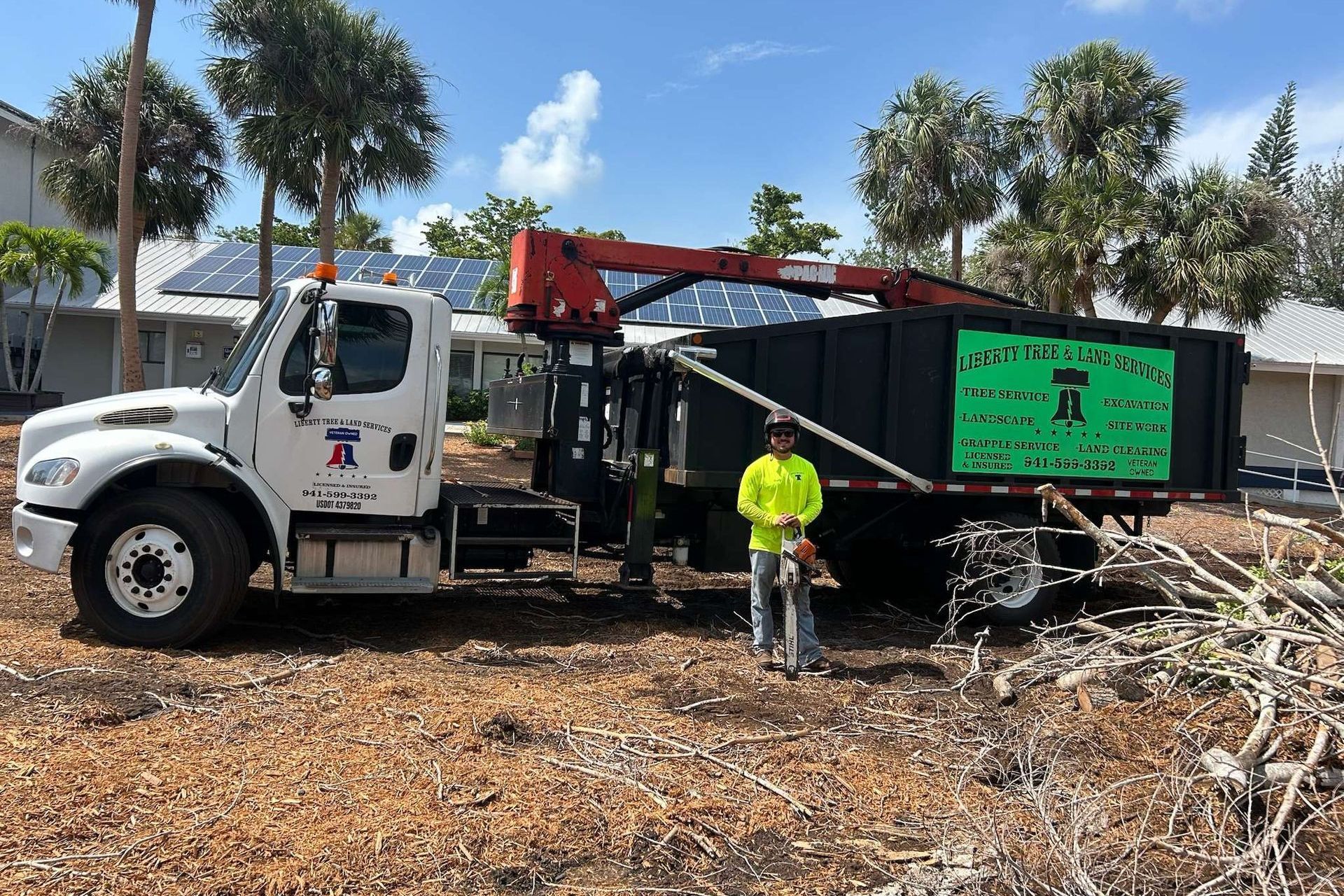 A tree service truck with a worker standing nearby. The truck has a crane arm and a full dumpster.