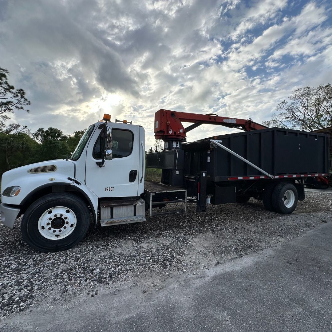 White truck with a crane and debris container parked on gravel. Cloudy sky in background.