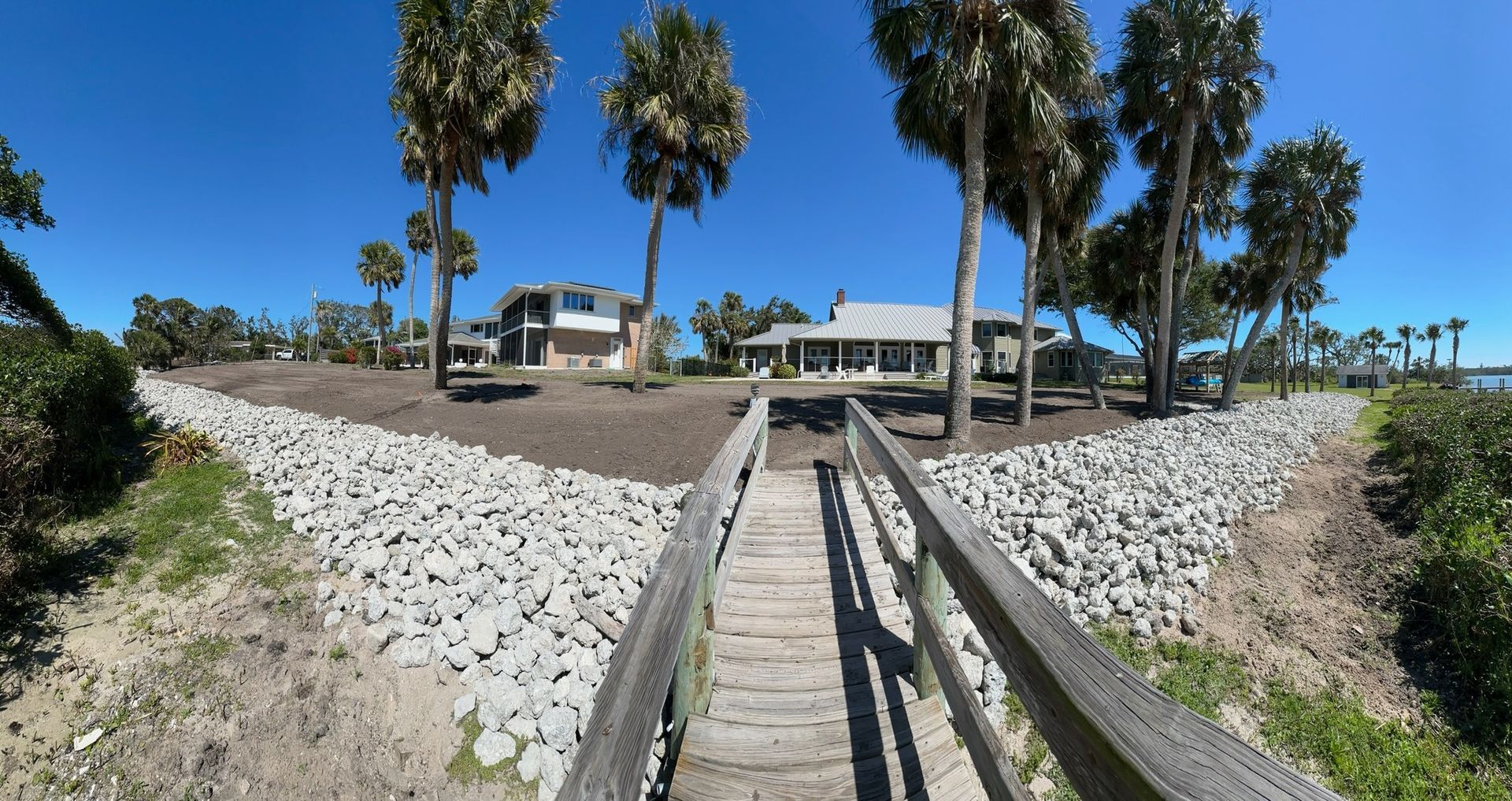 Wooden walkway between rock walls leads to waterfront houses under palm trees.