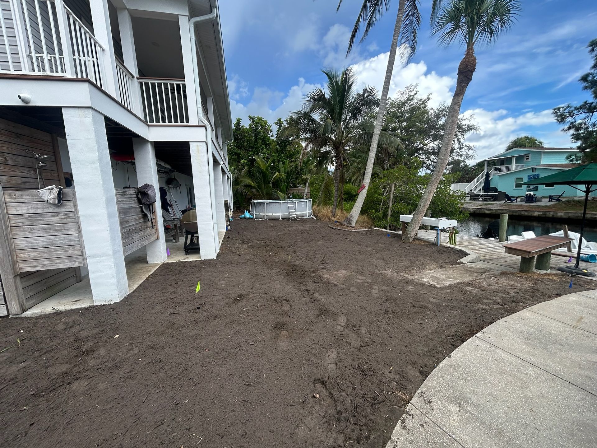 A cleared area of dark soil next to a white house, palm trees, and water.