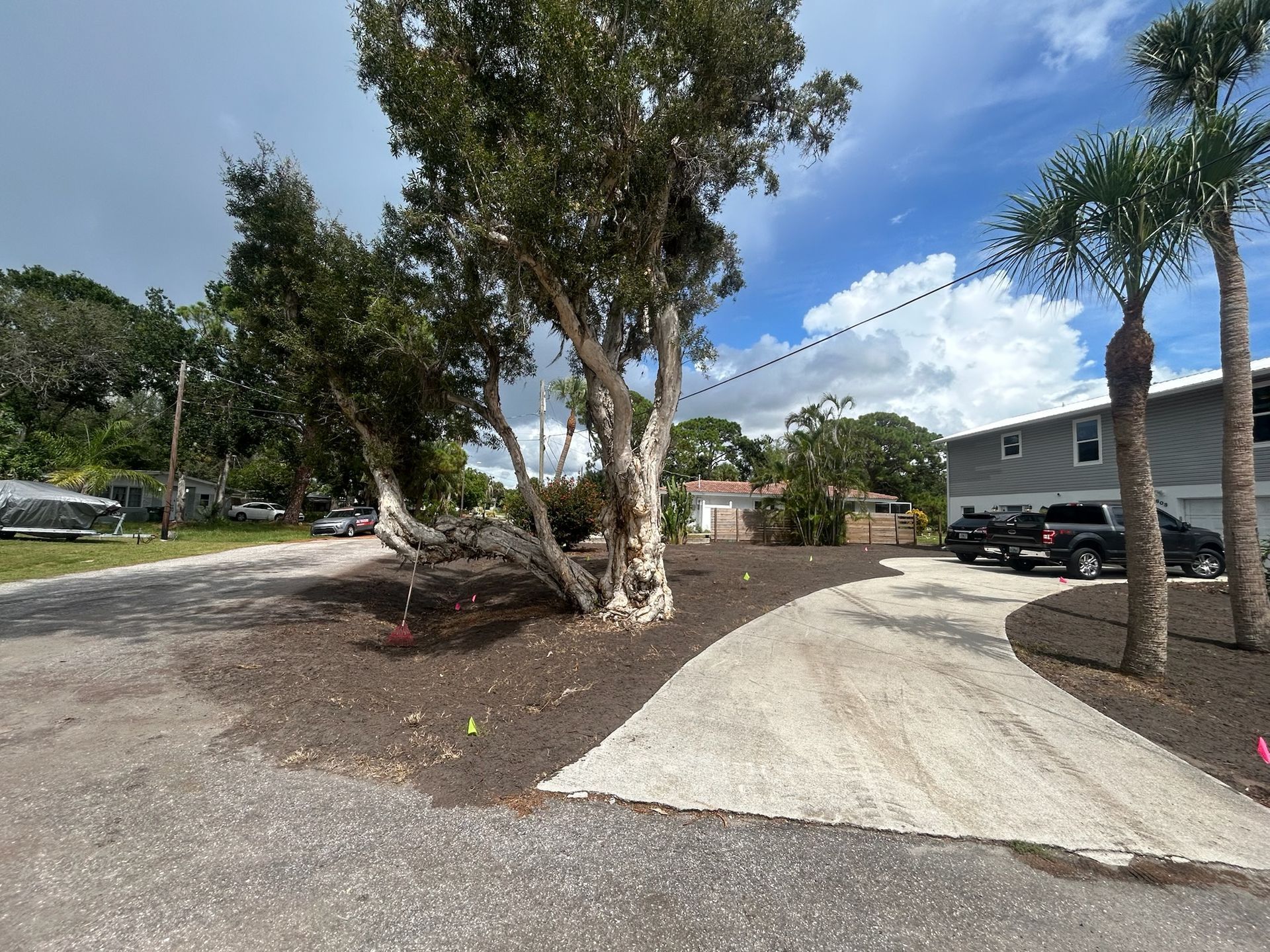 A gravel road leads to a driveway alongside a tree, with cars parked and a cloudy sky.