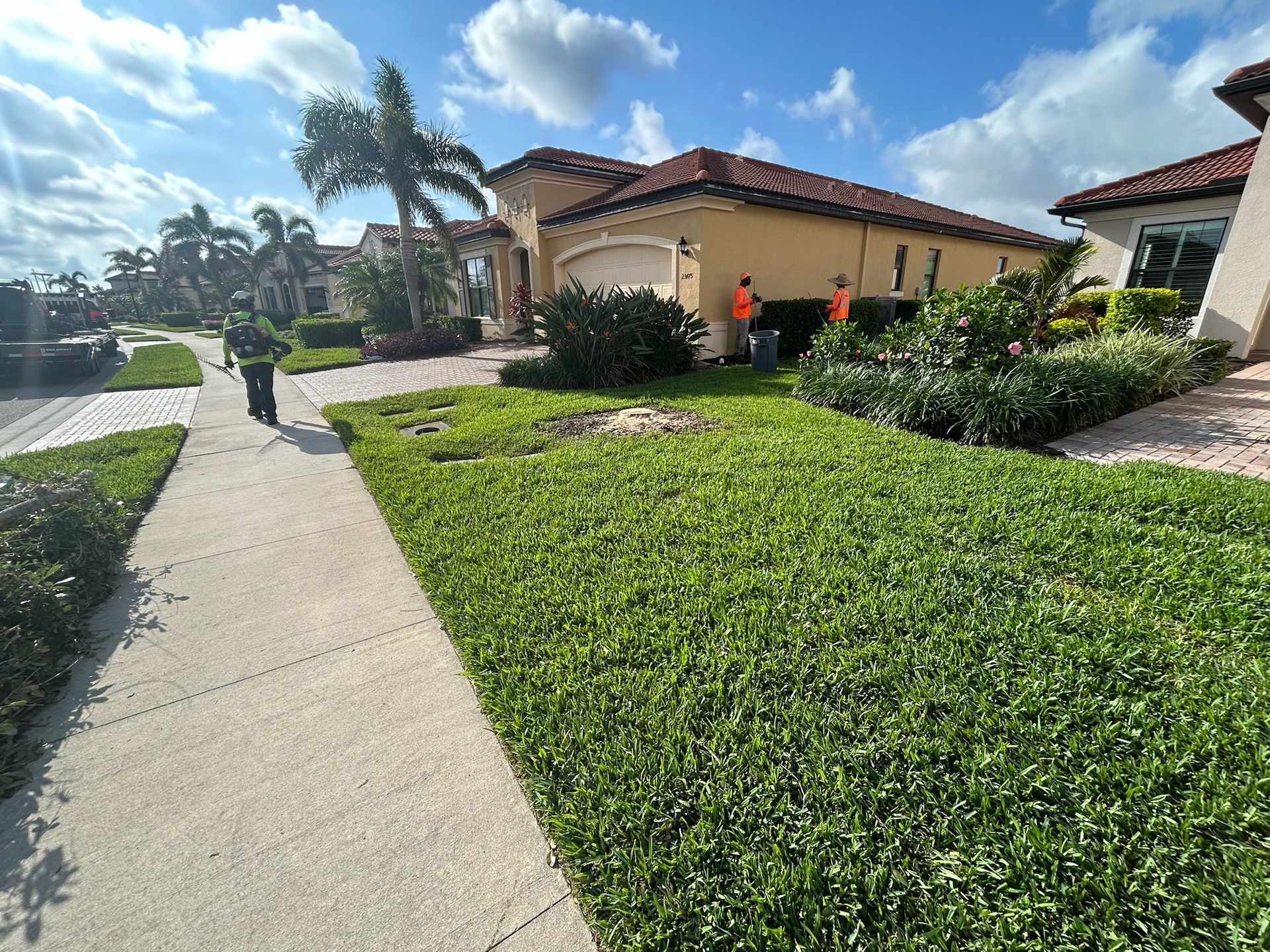 Green lawn and sidewalk in front of a house. Two workers near the garage are trimming bushes.