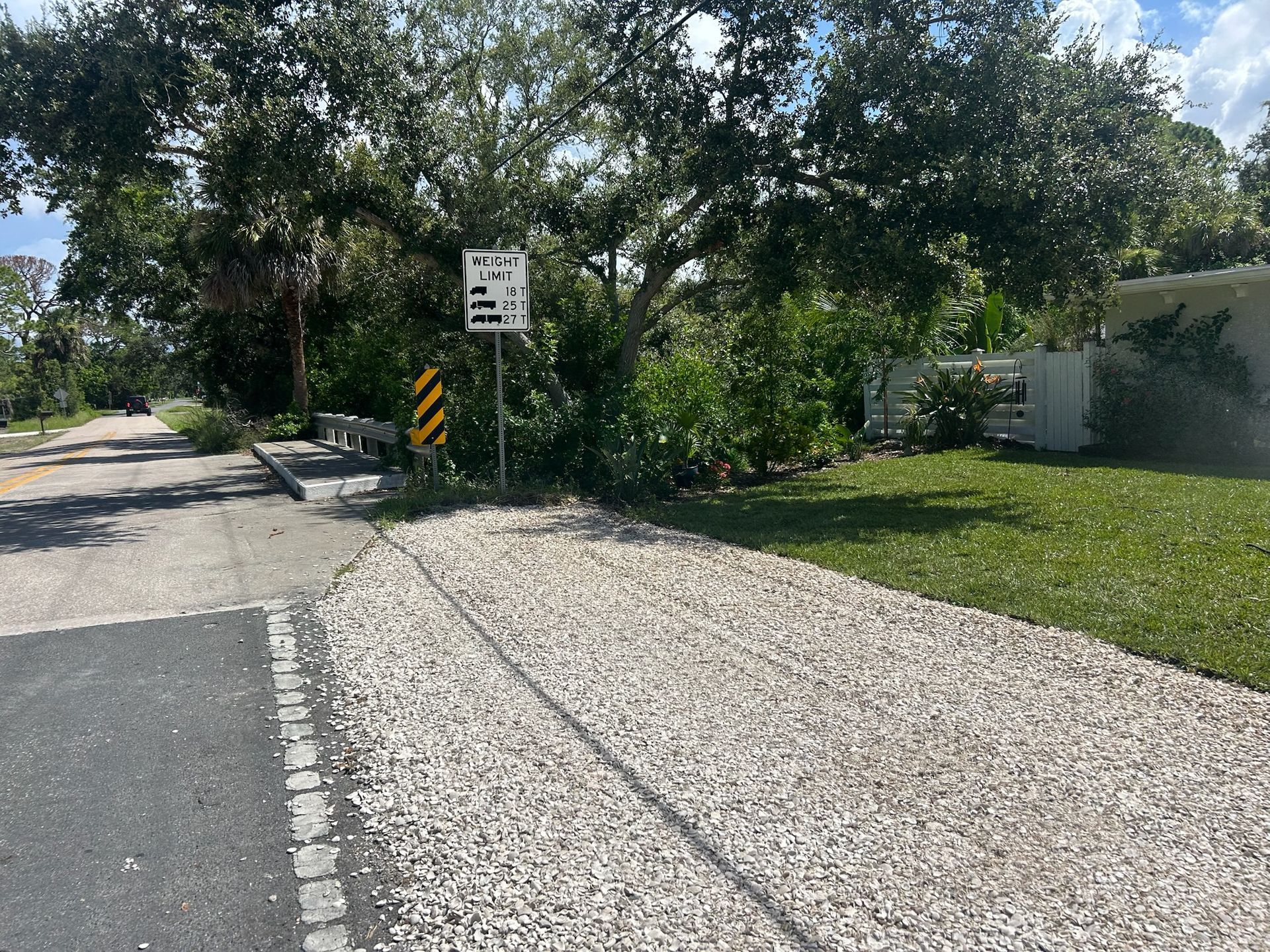 Gravel driveway leading to a paved road and a railroad crossing with signage, next to green grass and trees.