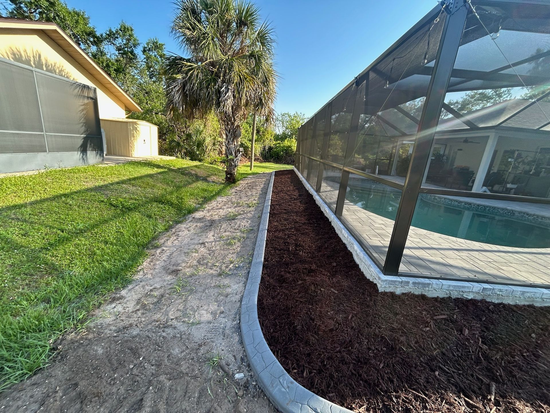 Poolside view: Brown mulch bed and concrete curb alongside a swimming pool enclosed in black screening; lush grass and a home.