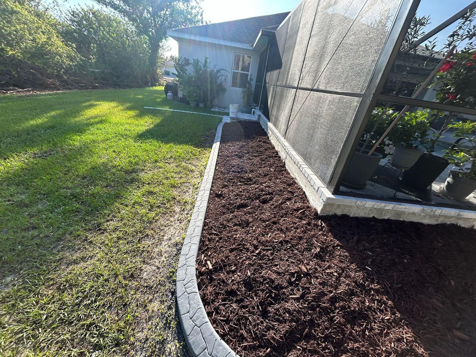 A garden bed with dark mulch, bordered by concrete pavers, next to a screened enclosure.