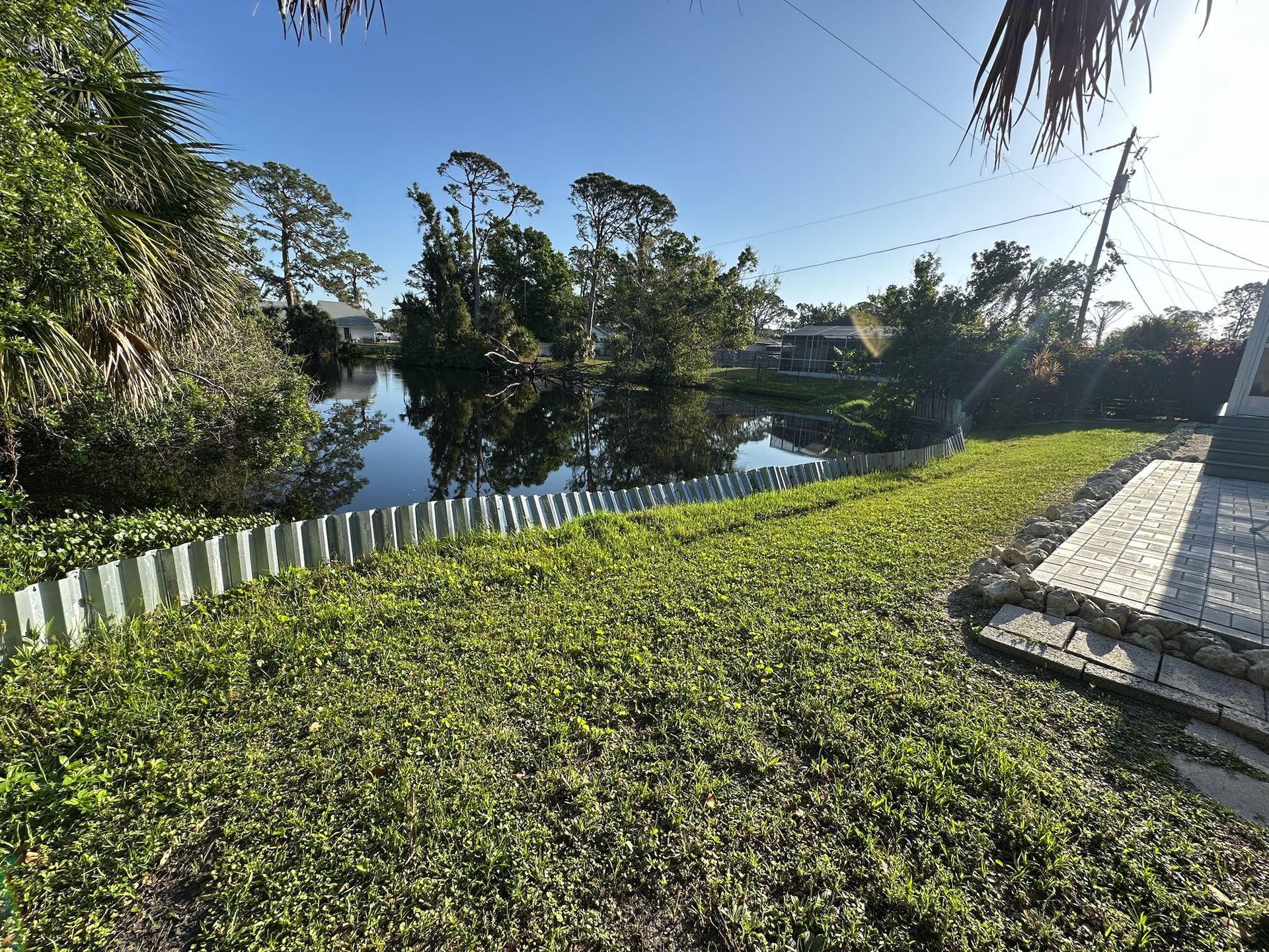 Grassy yard bordering a calm body of water, with trees in the background under a blue sky.