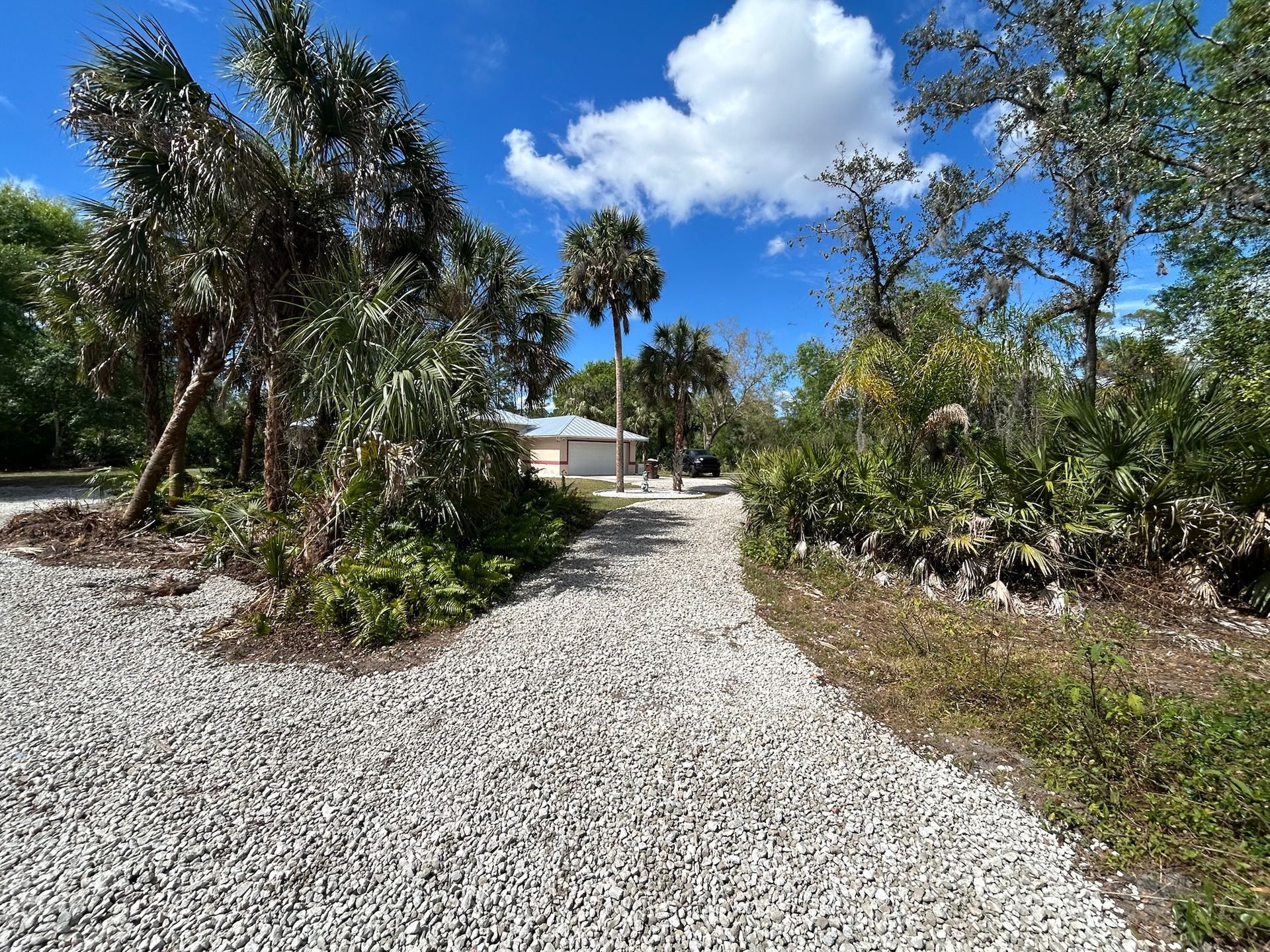 Gravel path leads toward a small building, surrounded by green foliage and palm trees, under a blue sky.