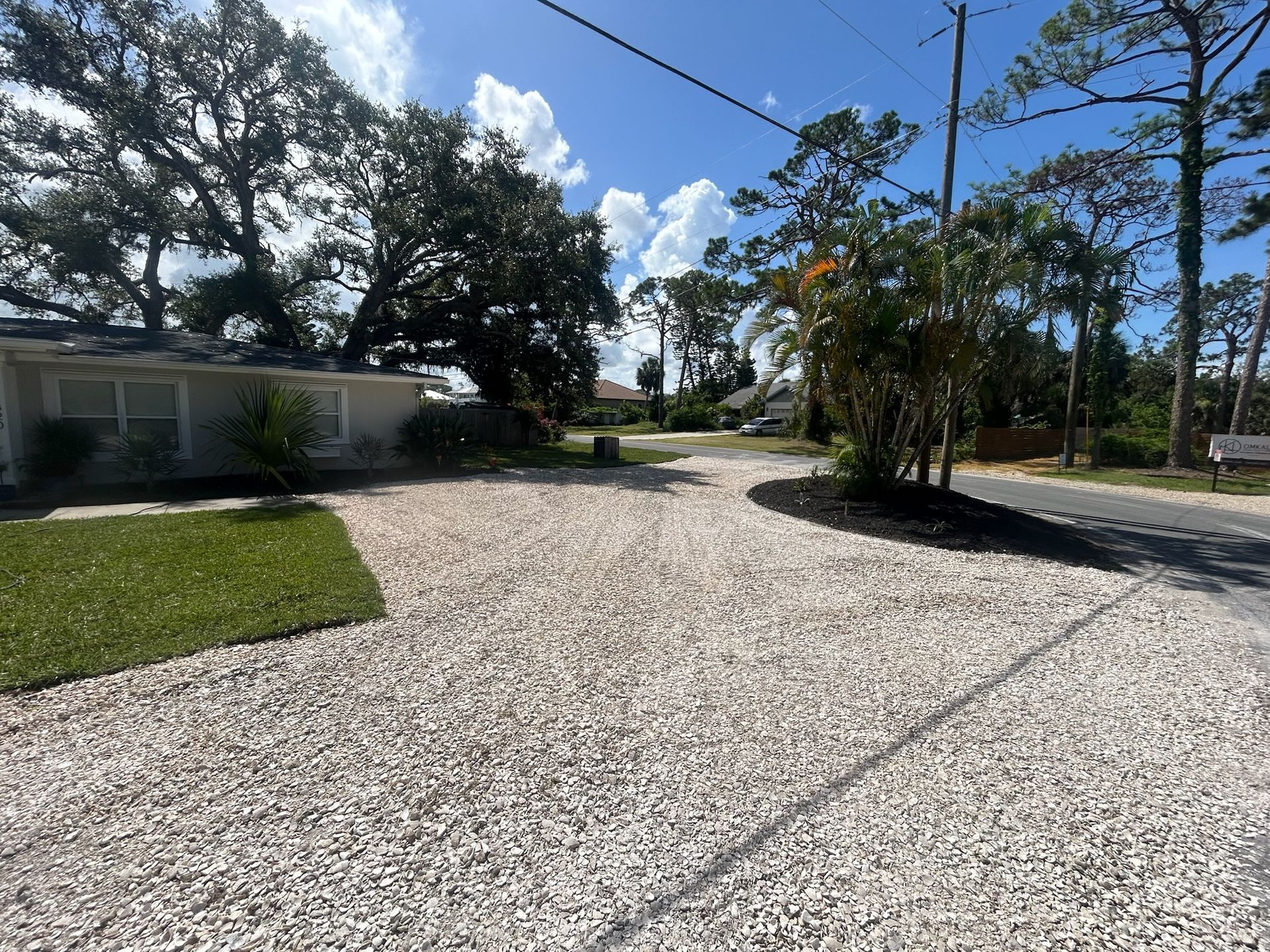 Gravel driveway leading to a house, flanked by grass and trees on a sunny day.