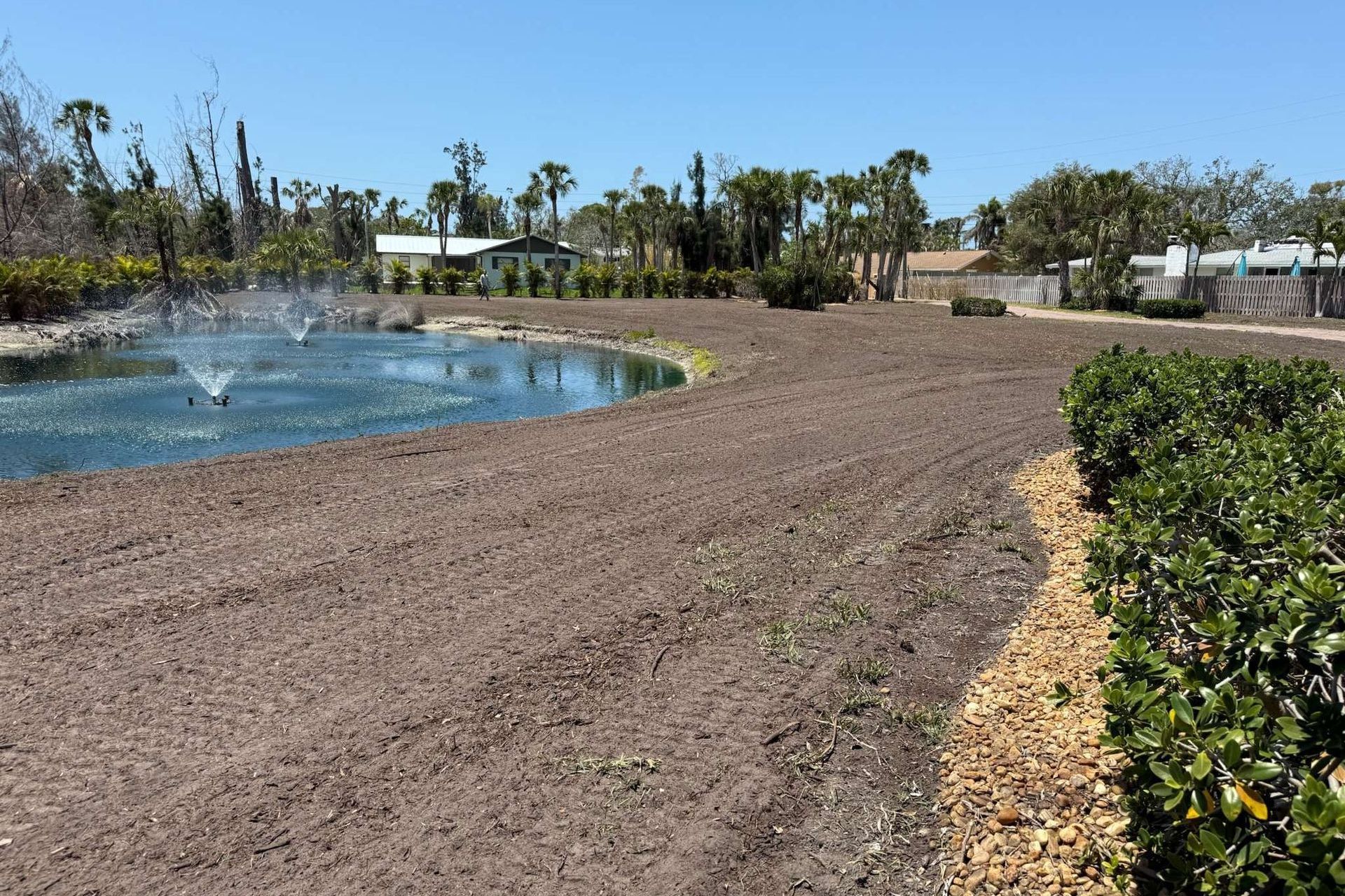 Pond with fountains, tilled brown earth, and shrubs under a clear blue sky, with houses in the background.