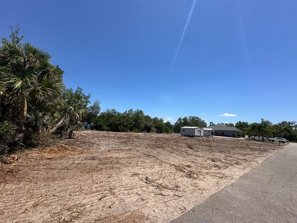Cleared land with some trees on the left and houses in the distance under a blue sky.