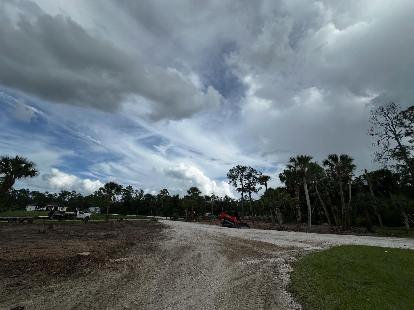Dirt road leads towards trees and cloudy sky. A small excavator sits in the distance.