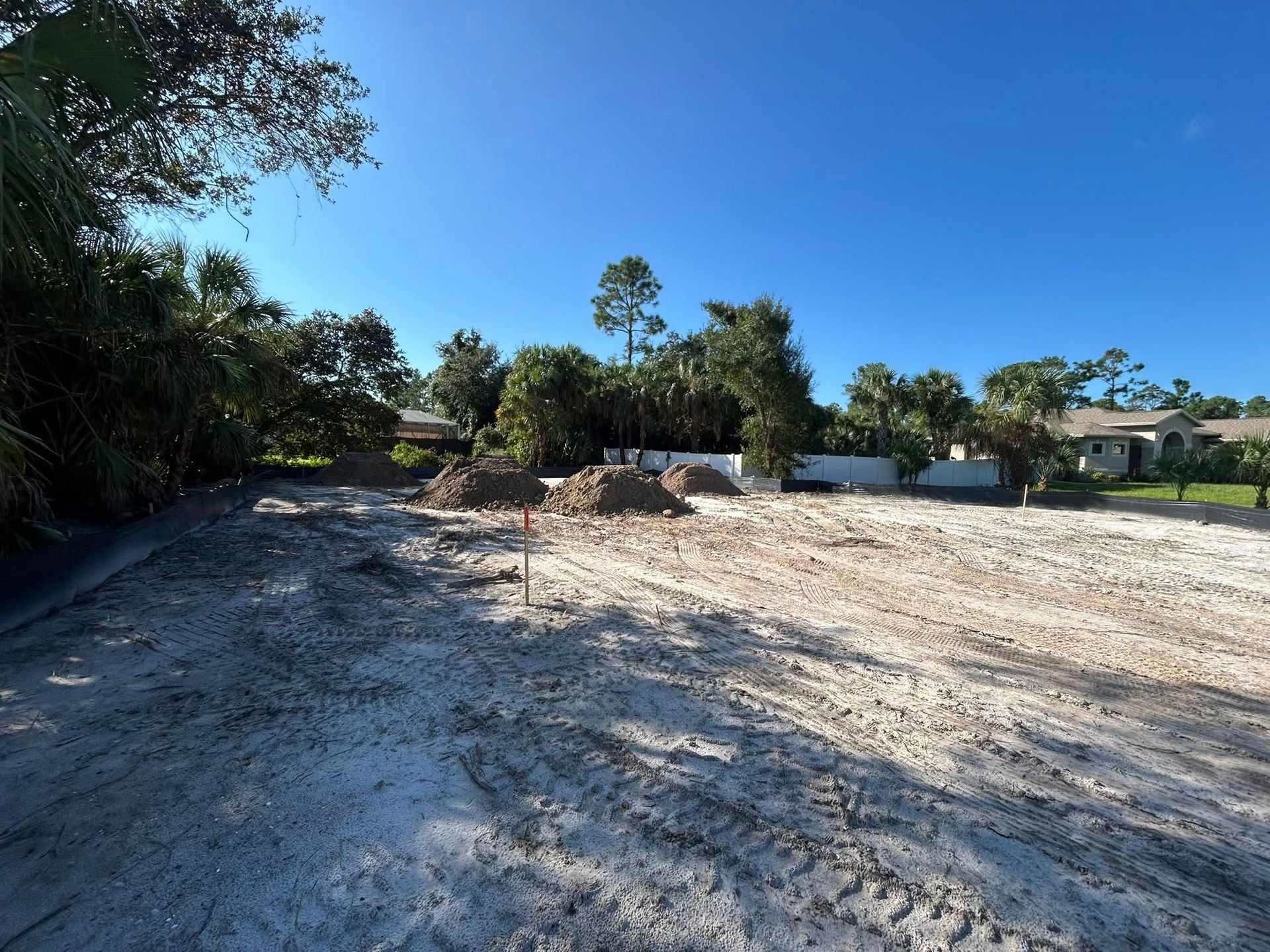 Cleared sandy lot with piles of mulch, trees, houses, and a clear blue sky.