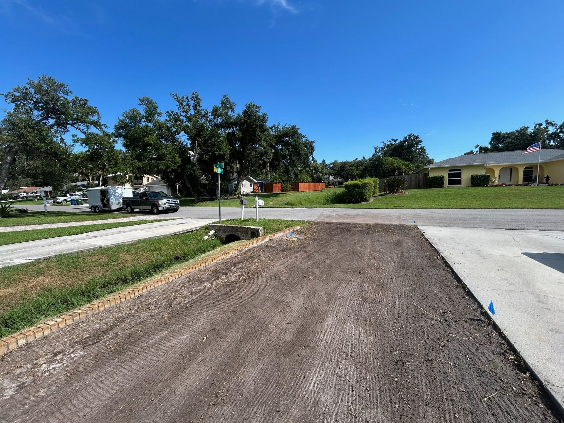 Driveway construction with dirt and gravel, street with houses, and blue sky.