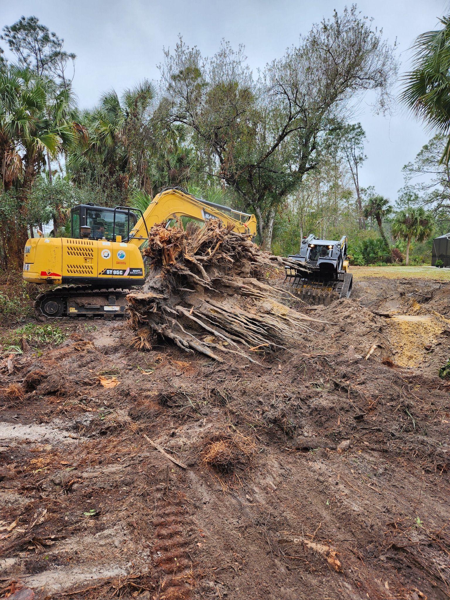 Yellow excavator and small white machine clearing a tree stump in a muddy area.