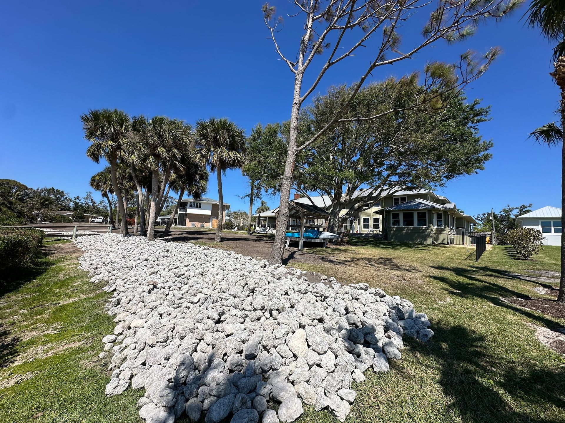 Rocks line a grassy embankment near a waterway, with trees and houses visible under a blue sky.