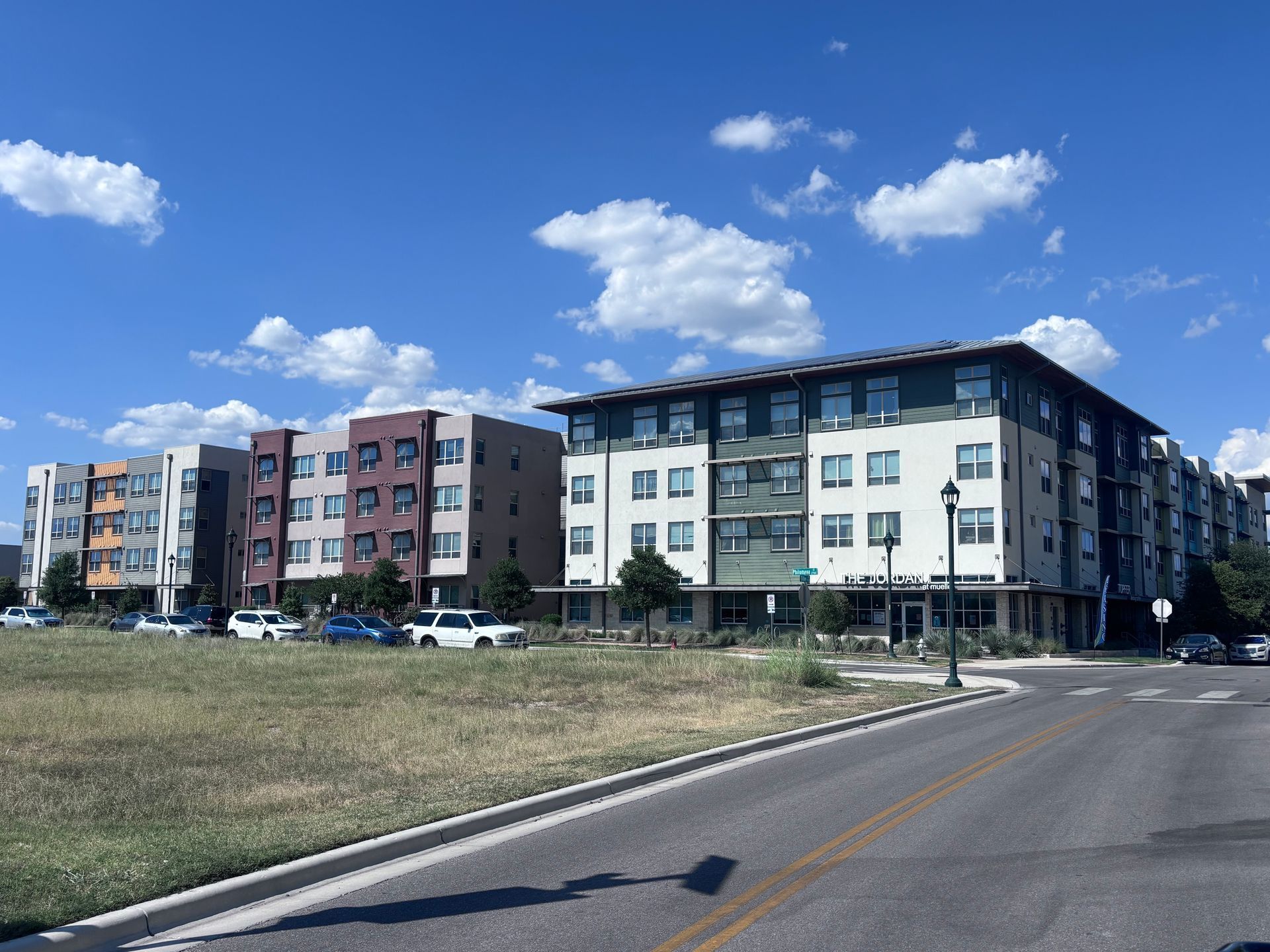 Apartment buildings with varying colors on a sunny day, near a grassy field and a road.