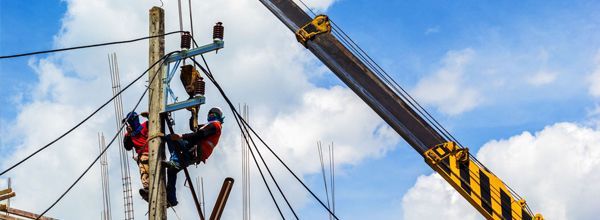 Linemen on a utility pole with a crane under a blue sky.
