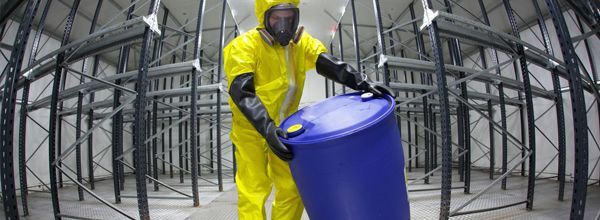 Person in yellow hazmat suit carrying blue barrel in a storage area with metal shelving.