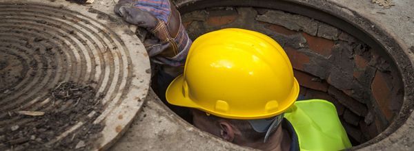 A worker wearing a yellow hard hat and safety vest, working in a manhole.