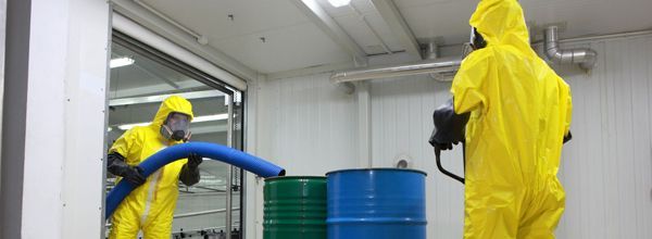 Two people in yellow hazmat suits vacuuming near barrels in a white room.