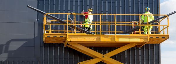Two workers in safety vests on a yellow scissor lift, working on a dark building.