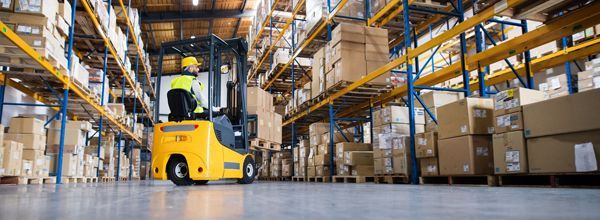 A yellow forklift in a warehouse with boxes on shelves. The driver is wearing a yellow vest.