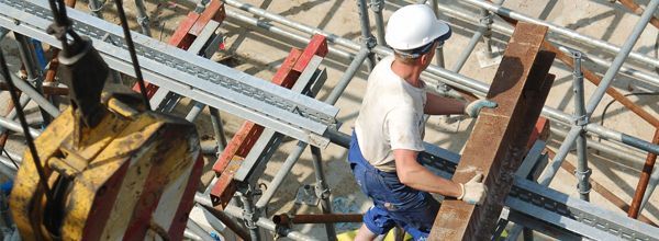 Construction worker carrying a long wooden beam on scaffolding, wearing a white hard hat and gloves.
