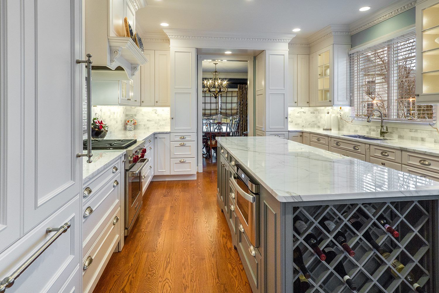Elegant white kitchen with a marble-topped island, wine rack, and stainless steel appliances.