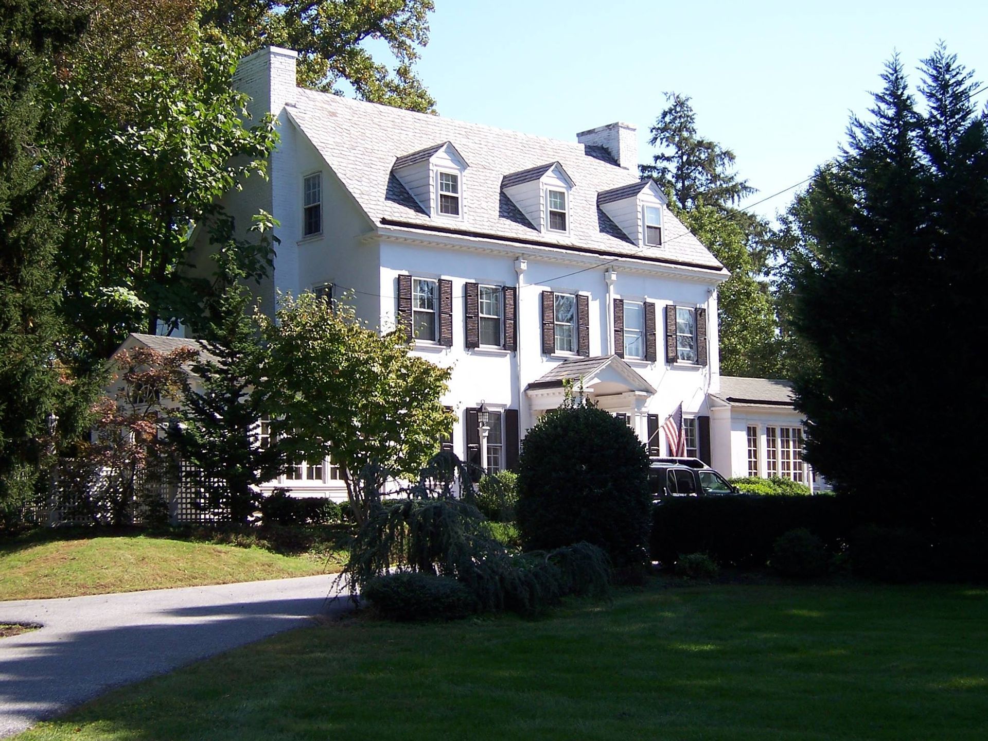 White two-story house with black shutters and a slate roof, set amidst trees and a green lawn.