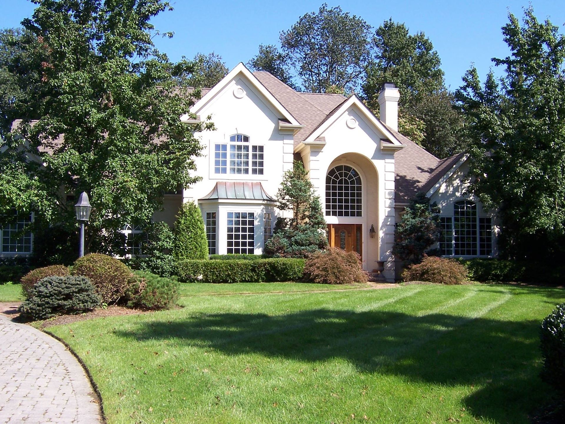 Cream-colored house with brown roof, arched entryway, lush green lawn, and trees; curved brick driveway.