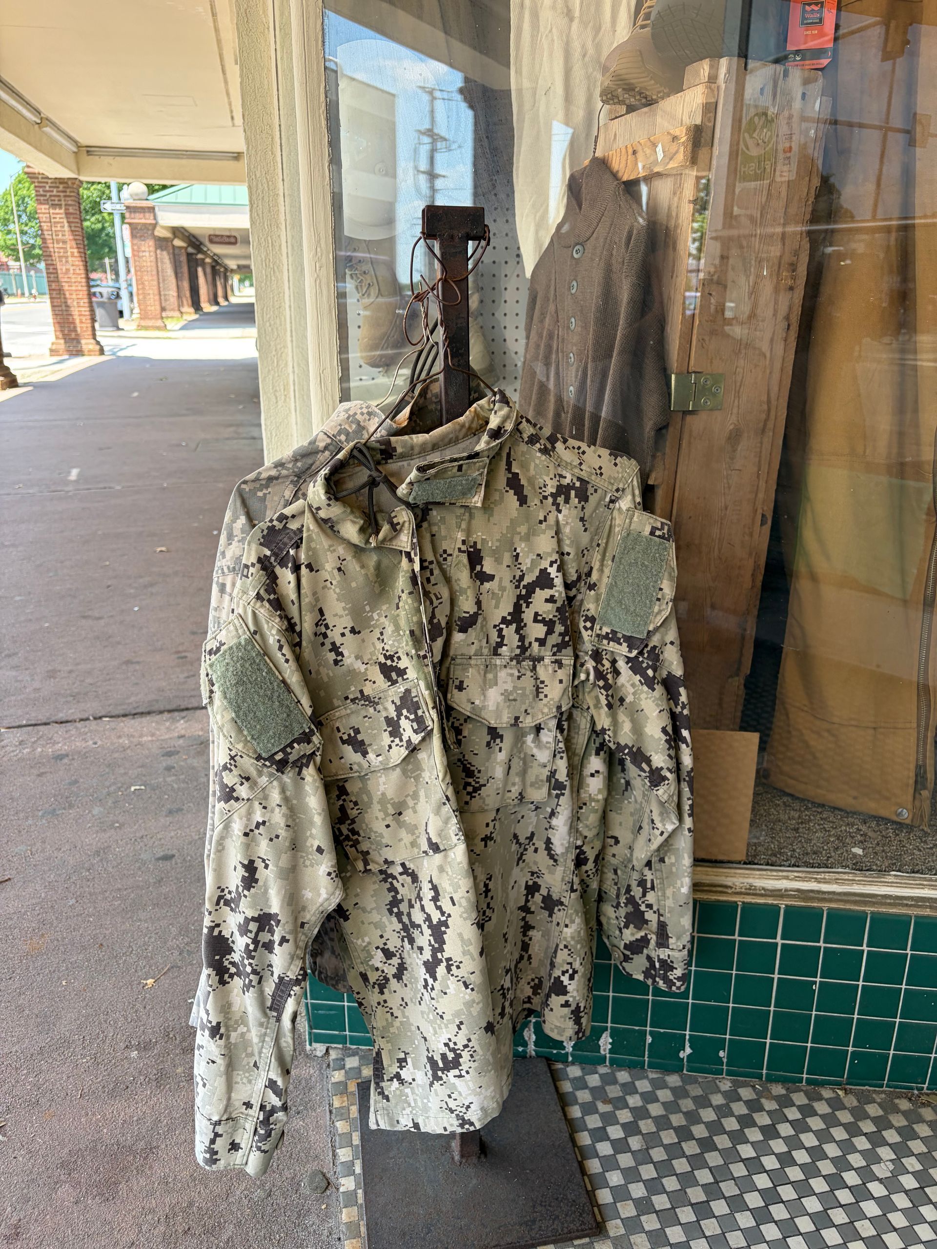 Camouflage jacket hanging outside a shop on a mannequin, beside a tiled wall and sidewalk.