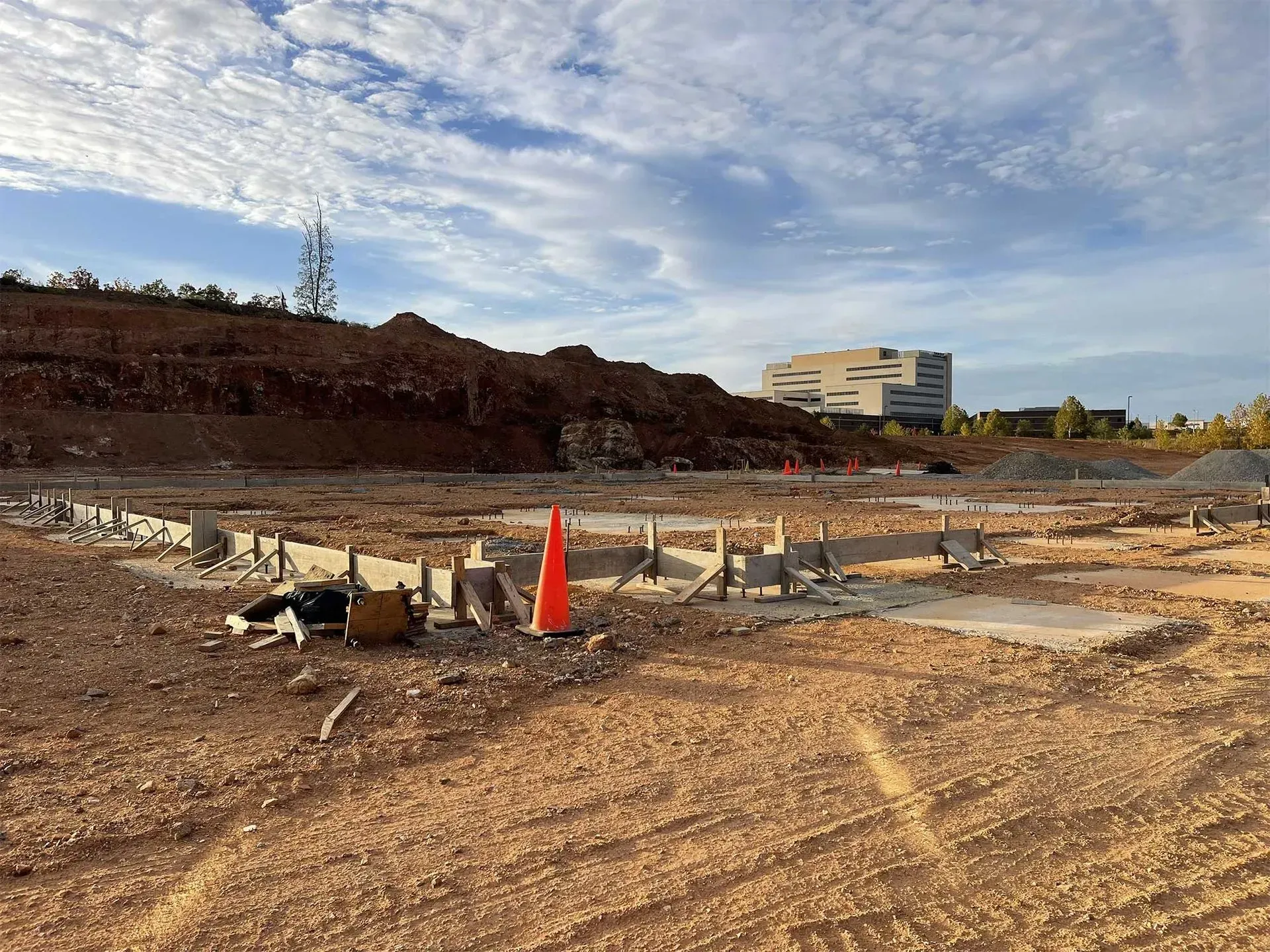 Construction site with foundation forms, dirt, and a building in the background under a cloudy sky.