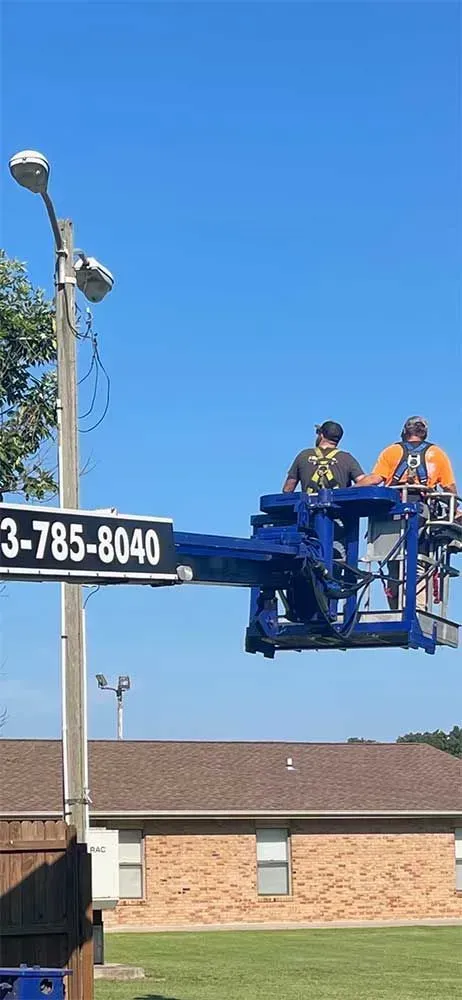 Two workers in a lift basket near a utility pole, bright blue sky.